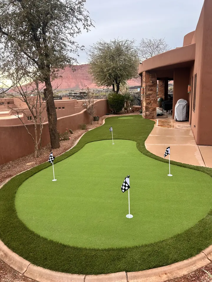 Finished backyard putting green with three checkered flags and a curved concrete curb beside an adobe style home, red rock behind