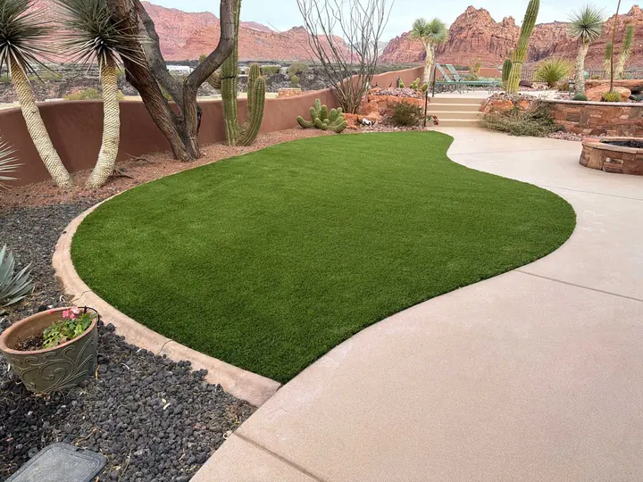 Curved artificial turf lawn beside a concrete patio with a fire pit, yuccas, cacti, and red rock cliffs