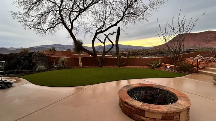 Curved artificial turf lawn beside a concrete patio with a round stone fire pit at twilight, red rock behind